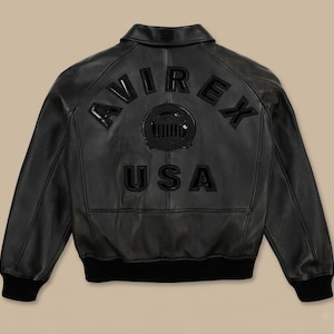 May include: Black leather jacket with the word "AVIREX" arched across the upper back, and "USA" below a circular emblem. The jacket has a collar, ribbed cuffs, and a ribbed hem. The jacket is a classic bomber style.