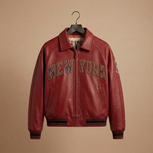 K&ouml;nnte beinhalten: Burgunderfarbene Lederjacke mit dem Schriftzug "NEW YORK" auf der Brust. Die Jacke hat einen Kragen, gerippte B&uuml;ndchen und einen gestreiften Saum. Sie h&auml;ngt an einem schwarzen Kleiderb&uuml;gel.