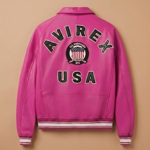 May include: A vibrant pink leather jacket with the word "AVIREX" arched across the shoulders. Below, a circular patch displays a US flag design and the text "ALL AMERICANS ESTABLISHED 1975". The word "USA" is printed below the patch. The jacket has white and burgundy striped cuffs.