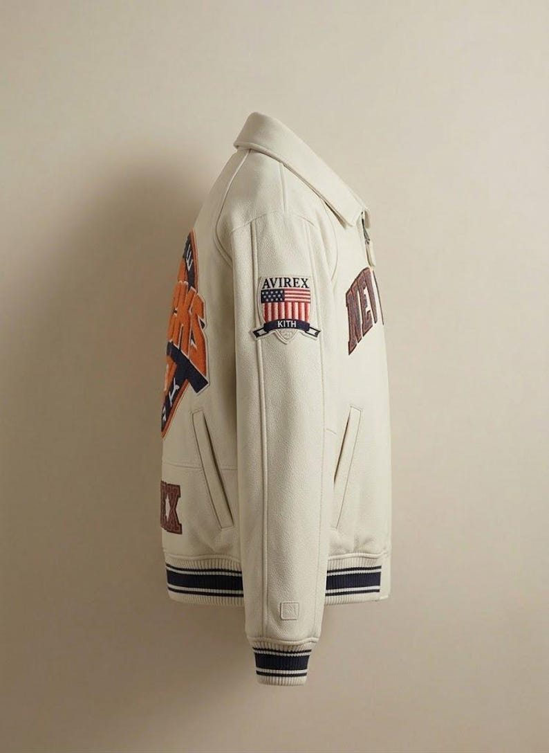 May include: Cream-colored varsity jacket with the text "NEW YORK" and "KITH" patches. The jacket features a navy and orange design on the back and sleeves, with a striped navy and white cuff and hem. The jacket has a collared neckline and a zipper closure.