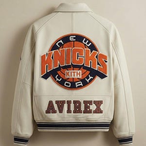 May include: Cream-colored leather jacket featuring the New York Knicks logo in orange and navy blue. The jacket has a ribbed hem and cuffs with navy and white stripes, and the brand name "AVIREX" is displayed below the logo.