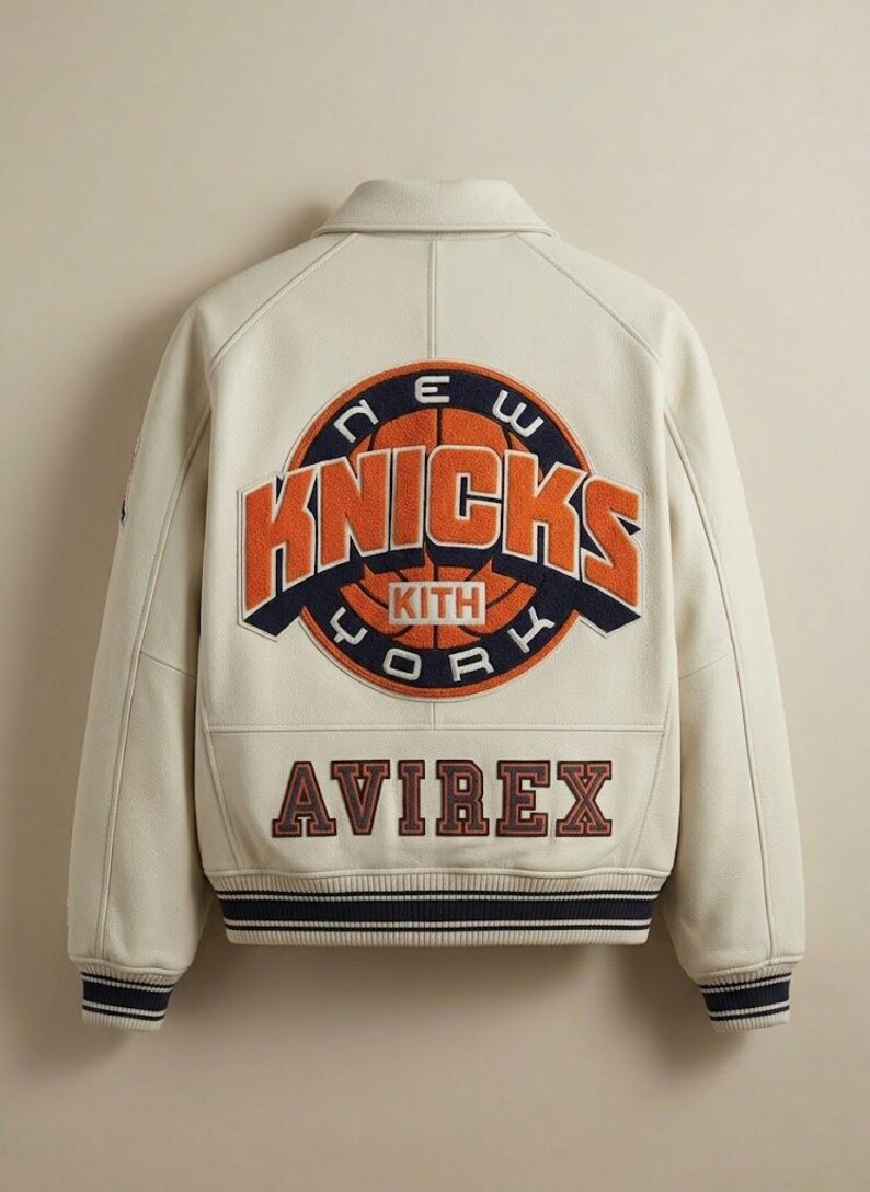 May include: Cream-colored leather jacket featuring the New York Knicks logo in orange and navy blue. The jacket has a ribbed hem and cuffs with navy and white stripes, and the brand name "AVIREX" is displayed below the logo.