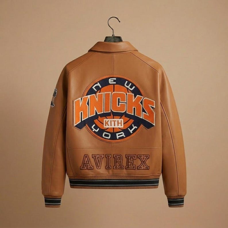 K&ouml;nnte beinhalten: Braune Lederjacke mit dem New York Knicks Logo auf dem R&uuml;cken. Die Jacke zeigt das Wort "Knicks" in Orange und Blau, mit "New York" dar&uuml;ber und "Kith" und "Avirex" darunter. Die Jacke hat einen Kragen und gestreifte Manschetten.