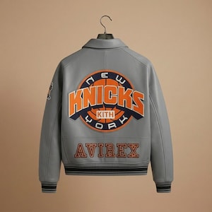 K&ouml;nnte beinhalten: Graue Lederjacke mit dem Logo der New York Knicks auf dem R&uuml;cken. Das Logo ist orange und blau, mit dem Wort "Knicks" prominent dargestellt. Die Jacke hat auch das "AVIREX"-Logo in Orange.