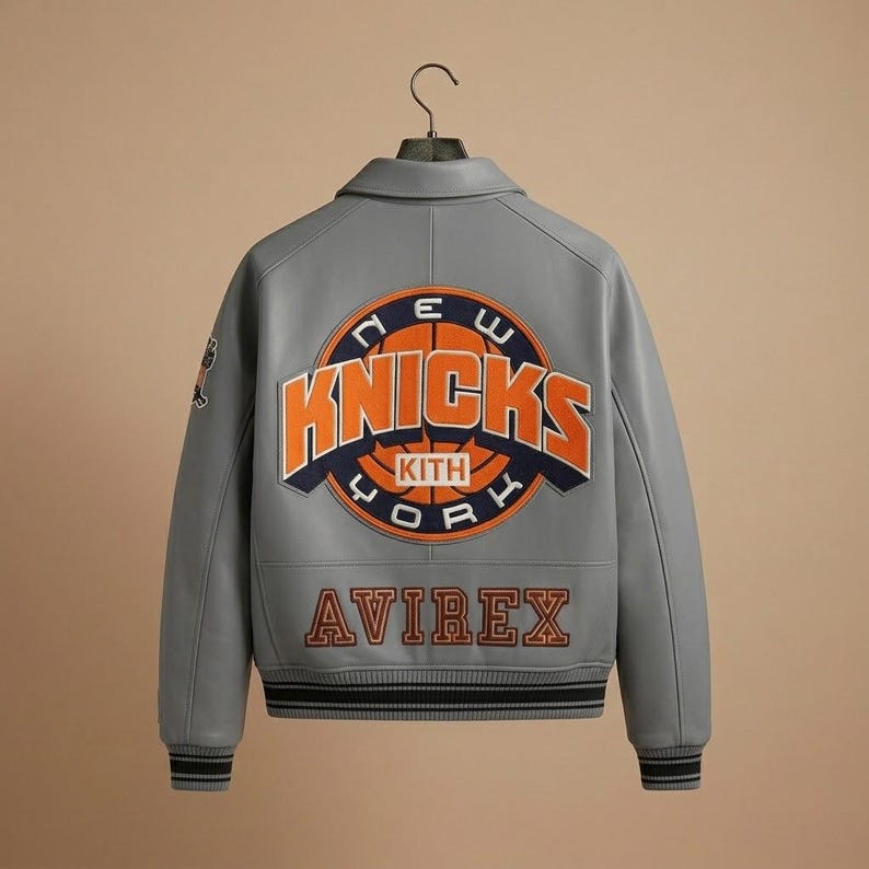 K&ouml;nnte beinhalten: Graue Lederjacke mit dem Logo der New York Knicks auf dem R&uuml;cken. Das Logo ist orange und blau, mit dem Wort "Knicks" prominent dargestellt. Die Jacke hat auch das "AVIREX"-Logo in Orange.