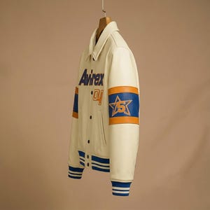 May include: An off-white varsity jacket featuring the text Avirex and the number 04 on the chest. The sleeves display blue and orange bands with a star graphic containing the number 5. The jacket has a collar, snap buttons, and striped cuffs.