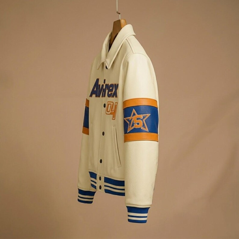 May include: An off-white varsity jacket featuring the text Avirex and the number 04 on the chest. The sleeves display blue and orange bands with a star graphic containing the number 5. The jacket has a collar, snap buttons, and striped cuffs.