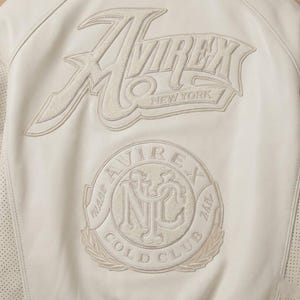May include: Cream-colored leather jacket with embroidered logos. The back features the brand name "Avirex New York" and a circular patch with "Cold Club" and "NYC" details. Perforated leather accents are visible.