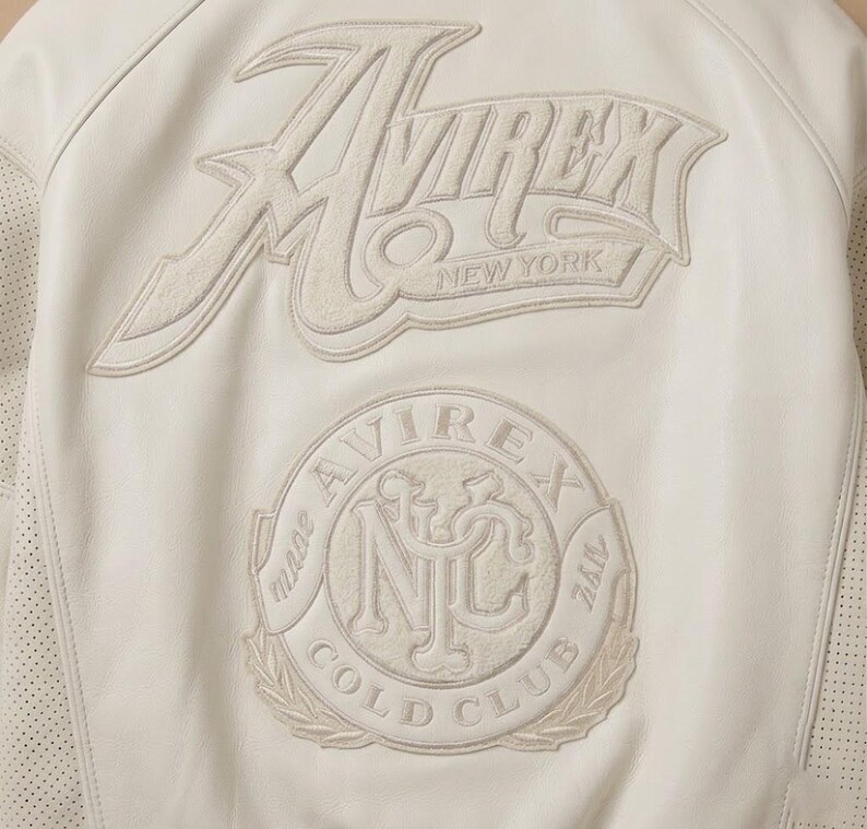 May include: Cream-colored leather jacket with embroidered logos. The back features the brand name "Avirex New York" and a circular patch with "Cold Club" and "NYC" details. Perforated leather accents are visible.