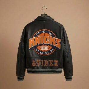 K&ouml;nnte beinhalten: Schwarze Lederjacke mit dem Logo der New York Knicks auf dem R&uuml;cken. Das Logo ist orange und blau, mit dem Wort "Knicks" prominent dargestellt. Die Jacke tr&auml;gt auch das Logo "Avirex".