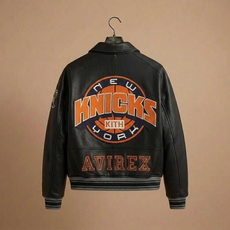 K&ouml;nnte beinhalten: Schwarze Lederjacke mit dem Logo der New York Knicks auf dem R&uuml;cken. Das Logo ist orange und blau, mit dem Wort "Knicks" prominent dargestellt. Die Jacke tr&auml;gt auch das Logo "Avirex".
