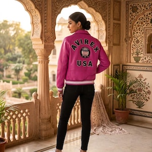 May include: A magenta leather jacket with the word "AVIREX" arched across the back, above a circular emblem and the word "USA". The jacket has a white and black striped trim on the cuffs and waist. The person is wearing black pants and white sneakers.
