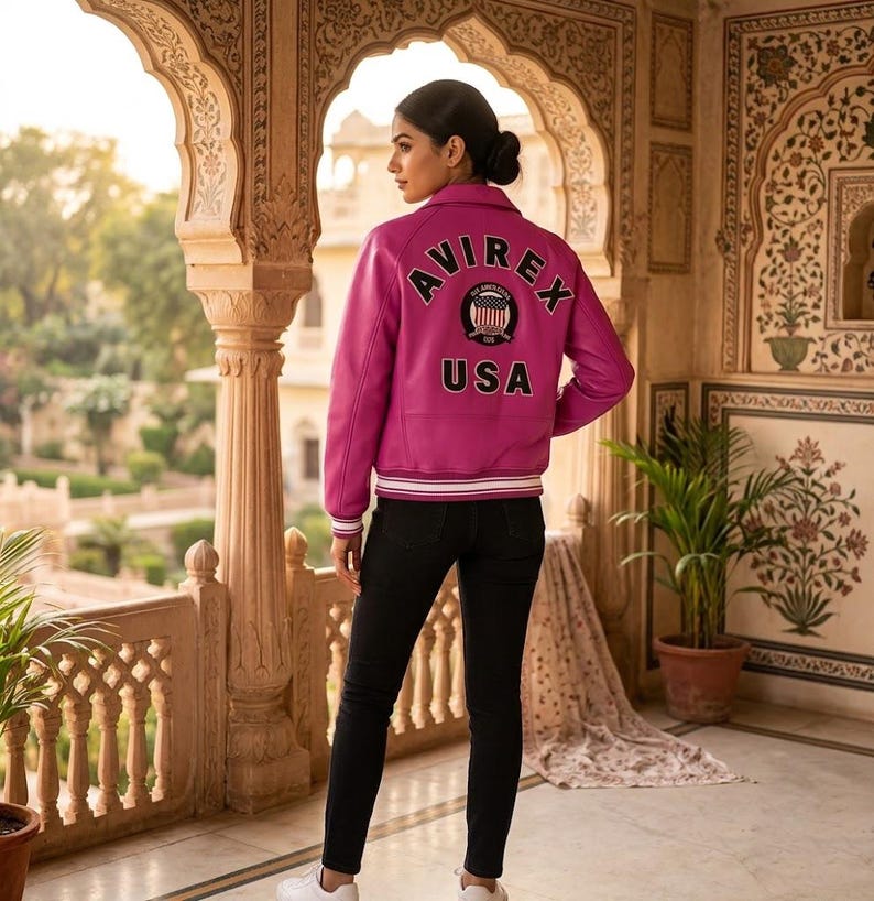 May include: A magenta leather jacket with the word "AVIREX" arched across the back, above a circular emblem and the word "USA". The jacket has a white and black striped trim on the cuffs and waist. The person is wearing black pants and white sneakers.