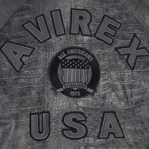 May include: A grey jacket with a textured crocodile skin pattern. The back features the text AVIREX USA in large grey letters. A circular patch in the center displays the text ALL AMERICANS ESTABLISHED 1975 and a shield with stars and stripes.