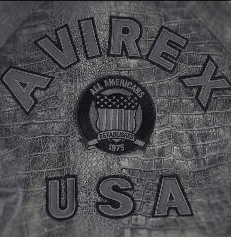 May include: A grey jacket with a textured crocodile skin pattern. The back features the text AVIREX USA in large grey letters. A circular patch in the center displays the text ALL AMERICANS ESTABLISHED 1975 and a shield with stars and stripes.
