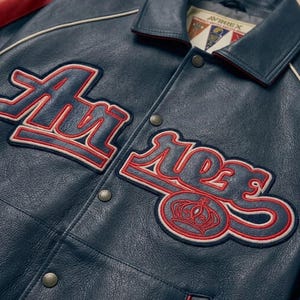 May include: Close-up of a navy blue leather jacket with red and white embroidered lettering and a crown emblem. The jacket features a collared neckline, snap closures, and the brand name "AVIREX" on a tag.