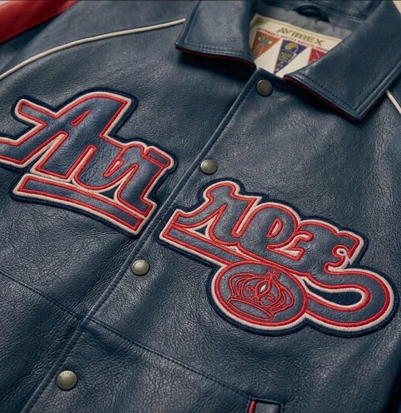 May include: Close-up of a navy blue leather jacket with red and white embroidered lettering and a crown emblem. The jacket features a collared neckline, snap closures, and the brand name "AVIREX" on a tag.