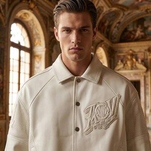 May include: Cream-colored leather jacket with a classic collar and button closure. Features a decorative emblem on the chest and perforated detailing on the sleeves. The jacket is a neutral color, suitable for various outfits.