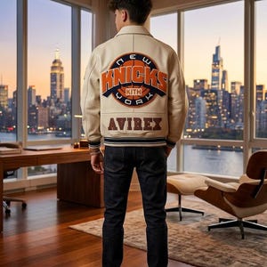 May include: Cream-colored leather jacket with the New York Knicks logo on the back, paired with dark wash jeans and white sneakers. The jacket features orange and blue accents, and the word "AVIREX" is displayed below the logo. The person is standing in front of a city skyline.