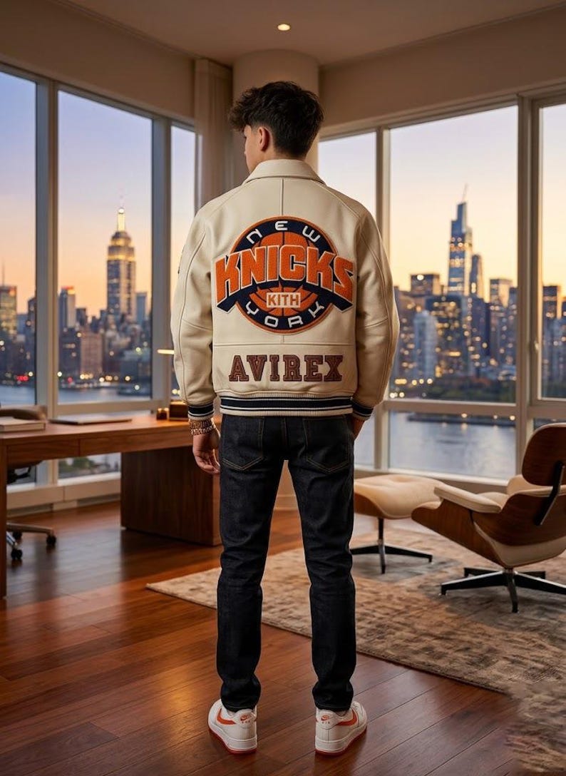 May include: Cream-colored leather jacket with the New York Knicks logo on the back, paired with dark wash jeans and white sneakers. The jacket features orange and blue accents, and the word "AVIREX" is displayed below the logo. The person is standing in front of a city skyline.