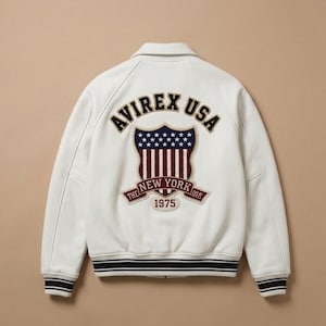 May include: A white varsity jacket featuring a large embroidered patch on the back. The patch displays the text AVIREX USA, a shield with stars and stripes, and a banner reading THE NEW YORK 1975. The jacket has a collar and striped ribbed cuffs.