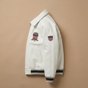 May include: A white jacket featuring a classic collar, a front zipper, and ribbed cuffs and hem with black and white stripes. The chest and sleeve display embroidered patches featuring a shield design with red, white, and blue American flag stripes.