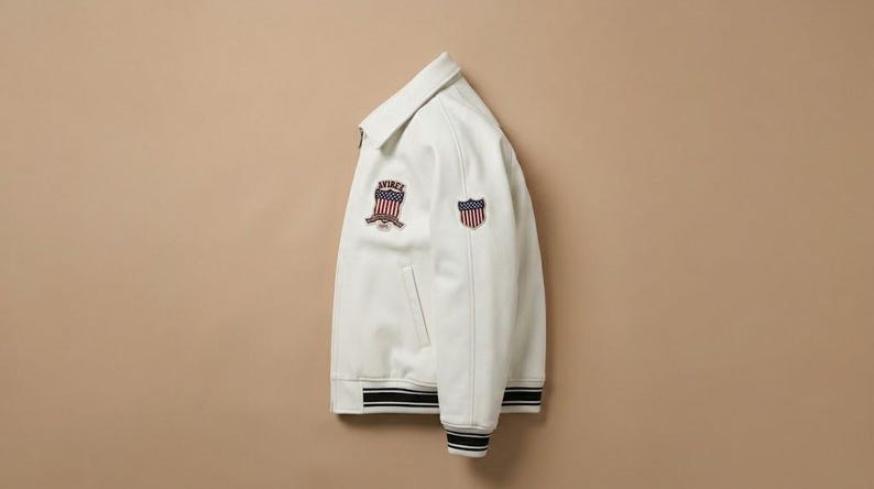 May include: A white jacket featuring a classic collar, a front zipper, and ribbed cuffs and hem with black and white stripes. The chest and sleeve display embroidered patches featuring a shield design with red, white, and blue American flag stripes.