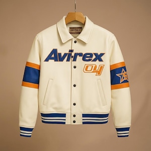 May include: An off-white varsity jacket featuring the text Avirex and 04 on the chest. The sleeves have blue and orange stripes with a star emblem on the left arm. It includes a pointed collar, snap button closure, and blue ribbed cuffs and hem.