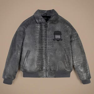 May include: A charcoal gray bomber jacket featuring a textured crocodile skin pattern throughout. The jacket has a front zipper closure, a standard collar, and ribbed knit cuffs and hem. A black embroidered patch with a shield design is on the chest.