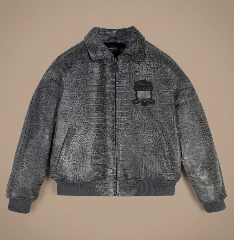 May include: A charcoal gray bomber jacket featuring a textured crocodile skin pattern throughout. The jacket has a front zipper closure, a standard collar, and ribbed knit cuffs and hem. A black embroidered patch with a shield design is on the chest.