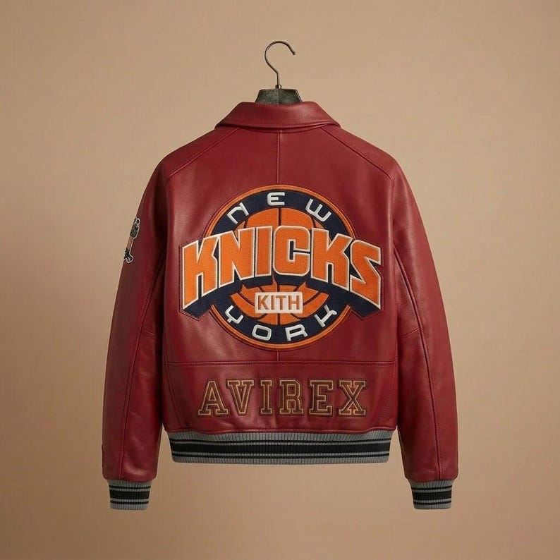 K&ouml;nnte beinhalten: Burgunderfarbene Lederjacke mit dem Logo der New York Knicks auf dem R&uuml;cken. Das Logo ist orange und blau, mit dem Wort "Knicks" prominent dargestellt. Die Jacke hat auch das "Avirex"-Logo und einen gestreiften Saum.