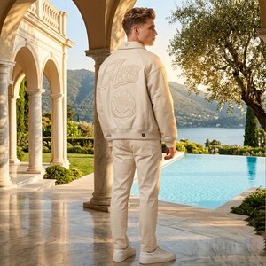 May include: A beige leather jacket with embroidered lettering and a circular emblem on the back, paired with matching beige pants and white sneakers. The outfit is worn by a person standing near a pool with a scenic background.