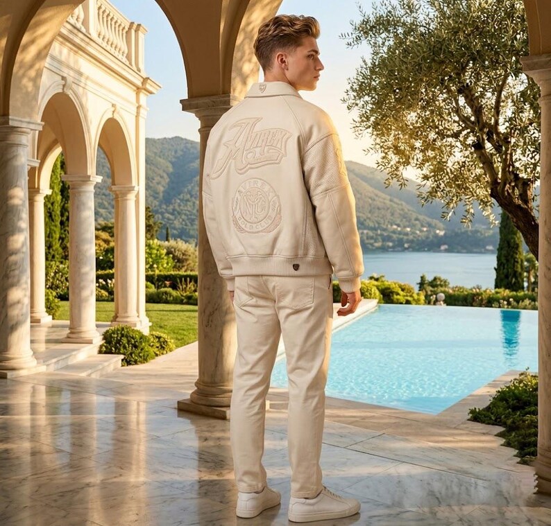 May include: A beige leather jacket with embroidered lettering and a circular emblem on the back, paired with matching beige pants and white sneakers. The outfit is worn by a person standing near a pool with a scenic background.