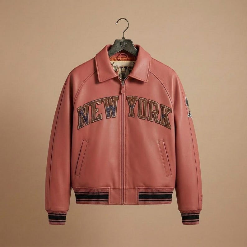 K&ouml;nnte beinhalten: Staubrosa Lederjacke mit dem Text "NEW YORK" auf der Brust. Die Jacke hat einen Kragen, einen Rei&szlig;verschluss und zwei Vordertaschen. Die Manschetten und der Saum weisen schwarz-marineblaue Streifen auf.