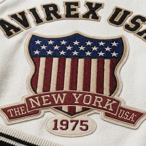May include: An off-white varsity jacket featuring a large embroidered patch on the back. The patch displays the text AVIREX USA above a shield with stars and stripes, and a banner reading THE NEW YORK U.S.A. with the year 1975 below it.