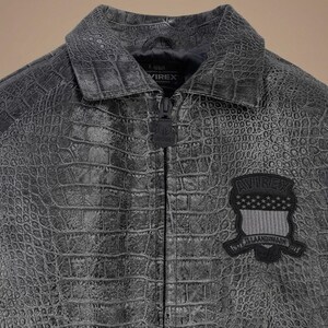 May include: A charcoal gray jacket with a textured crocodile skin pattern, featuring a pointed collar, a central zipper with a branded pull, and a black patch on the chest displaying the text AVIREX above a shield with stars and stripes.