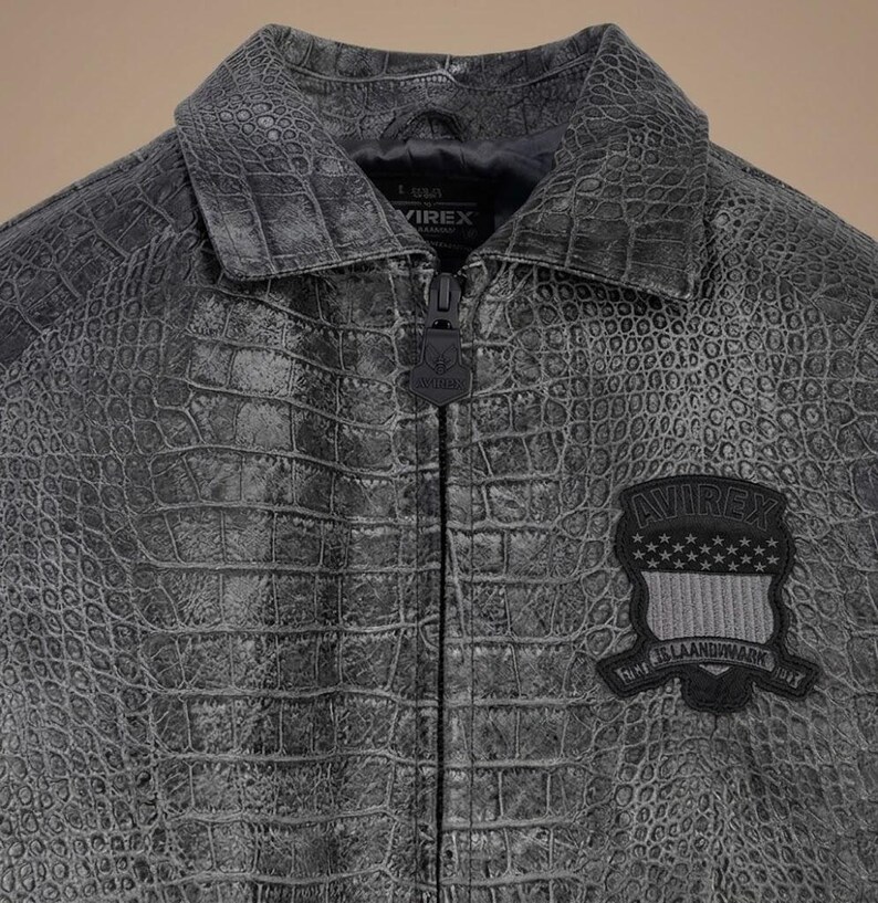May include: A charcoal gray jacket with a textured crocodile skin pattern, featuring a pointed collar, a central zipper with a branded pull, and a black patch on the chest displaying the text AVIREX above a shield with stars and stripes.