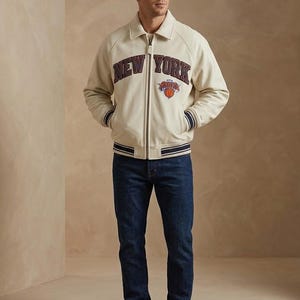 May include: Cream-colored bomber jacket with "NEW YORK" lettering and a basketball logo. The jacket has navy blue and white striped cuffs and a matching collar. The person is wearing dark blue jeans and white sneakers.