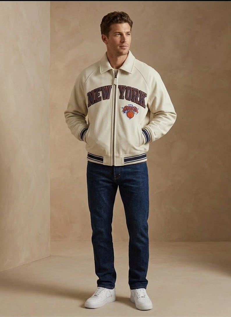 May include: Cream-colored bomber jacket with "NEW YORK" lettering and a basketball logo. The jacket has navy blue and white striped cuffs and a matching collar. The person is wearing dark blue jeans and white sneakers.