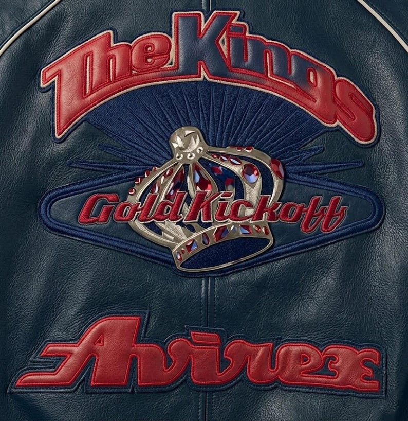 May include: Close-up of a navy blue leather jacket with red and gold embroidered details. The jacket features the text "The Kings", "Gold Kickoff", and "Avirex", along with a silver crown emblem.