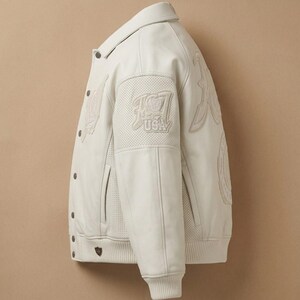 May include: Cream-colored leather jacket with a button-up front, collar, and ribbed cuffs. Features embroidered details and the text "USA" on the upper left sleeve. The jacket has a classic varsity style.