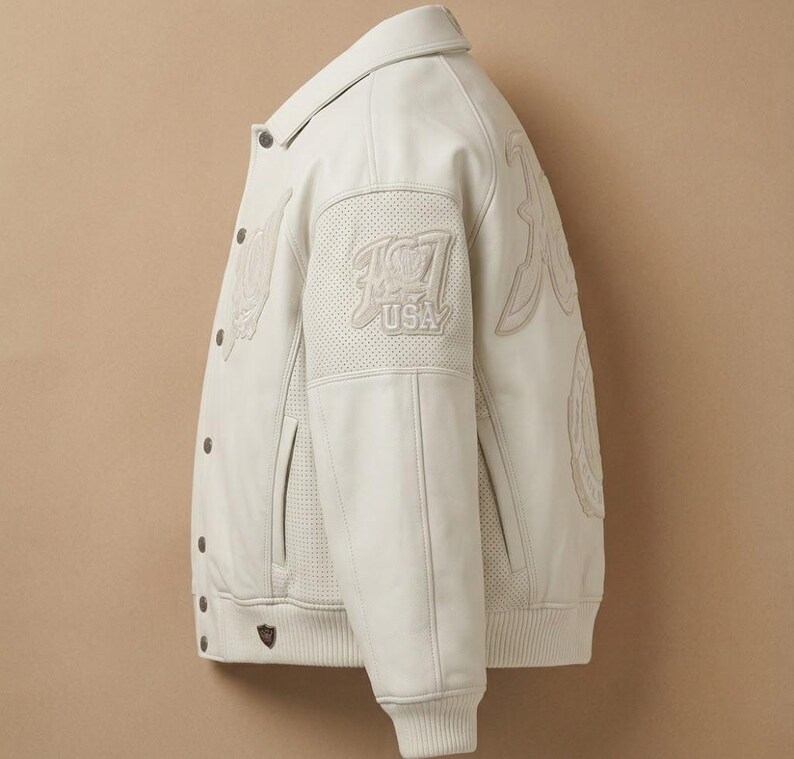 May include: Cream-colored leather jacket with a button-up front, collar, and ribbed cuffs. Features embroidered details and the text "USA" on the upper left sleeve. The jacket has a classic varsity style.