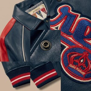 May include: A navy blue leather jacket with red and white accents. Features include a red shoulder panel, white piping, and a navy blue ribbed cuff with red and white stripes. The jacket has a patch that says "Gold Kickoff".