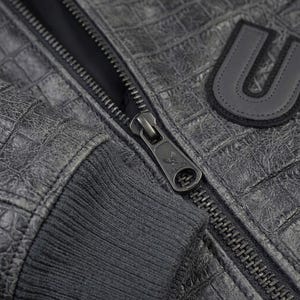May include: A dark gray jacket with a textured crocodile skin pattern, featuring a ribbed cuff and a metal zipper pull embossed with an eagle logo and the text AVIREX. A black letter U patch with a stitched border is visible on the chest area.