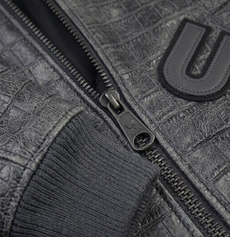 May include: A dark gray jacket with a textured crocodile skin pattern, featuring a ribbed cuff and a metal zipper pull embossed with an eagle logo and the text AVIREX. A black letter U patch with a stitched border is visible on the chest area.