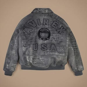 May include: A grey leather bomber jacket with a textured crocodile skin pattern. The back features the text AVIREX in an arch, the word USA below it, and a circular patch in the center with the text ALL AMERICANS ESTABLISHED 1975.