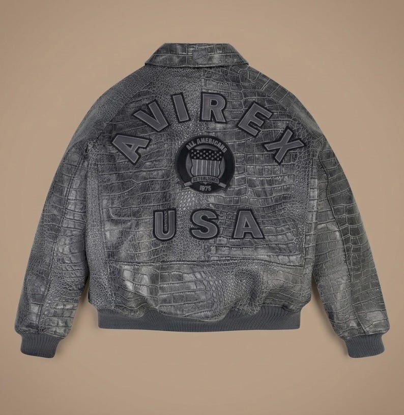 May include: A grey leather bomber jacket with a textured crocodile skin pattern. The back features the text AVIREX in an arch, the word USA below it, and a circular patch in the center with the text ALL AMERICANS ESTABLISHED 1975.