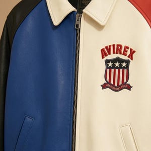 Peut inclure: Une veste en cuir color-block avec des manches noires, un panneau avant bleu et un panneau avant cr&egrave;me avec un col. La poitrine pr&eacute;sente un &eacute;cusson brod&eacute; AVIREX avec un bouclier, des &eacute;toiles, des rayures et le texte LIBERCIUNNANT PURIA.