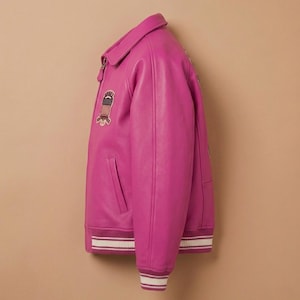 May include: A vibrant fuchsia leather jacket with a classic varsity style. Features a collared neckline, a front zipper, and a patch with an American flag design. The cuffs and hem have white and burgundy striped detailing.