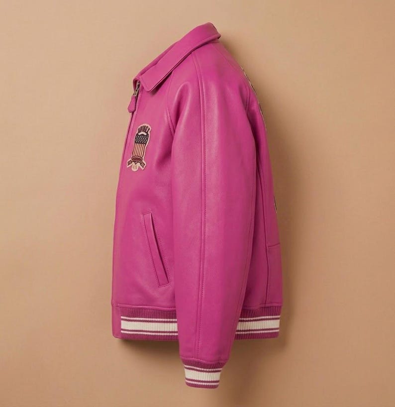 May include: A vibrant fuchsia leather jacket with a classic varsity style. Features a collared neckline, a front zipper, and a patch with an American flag design. The cuffs and hem have white and burgundy striped detailing.